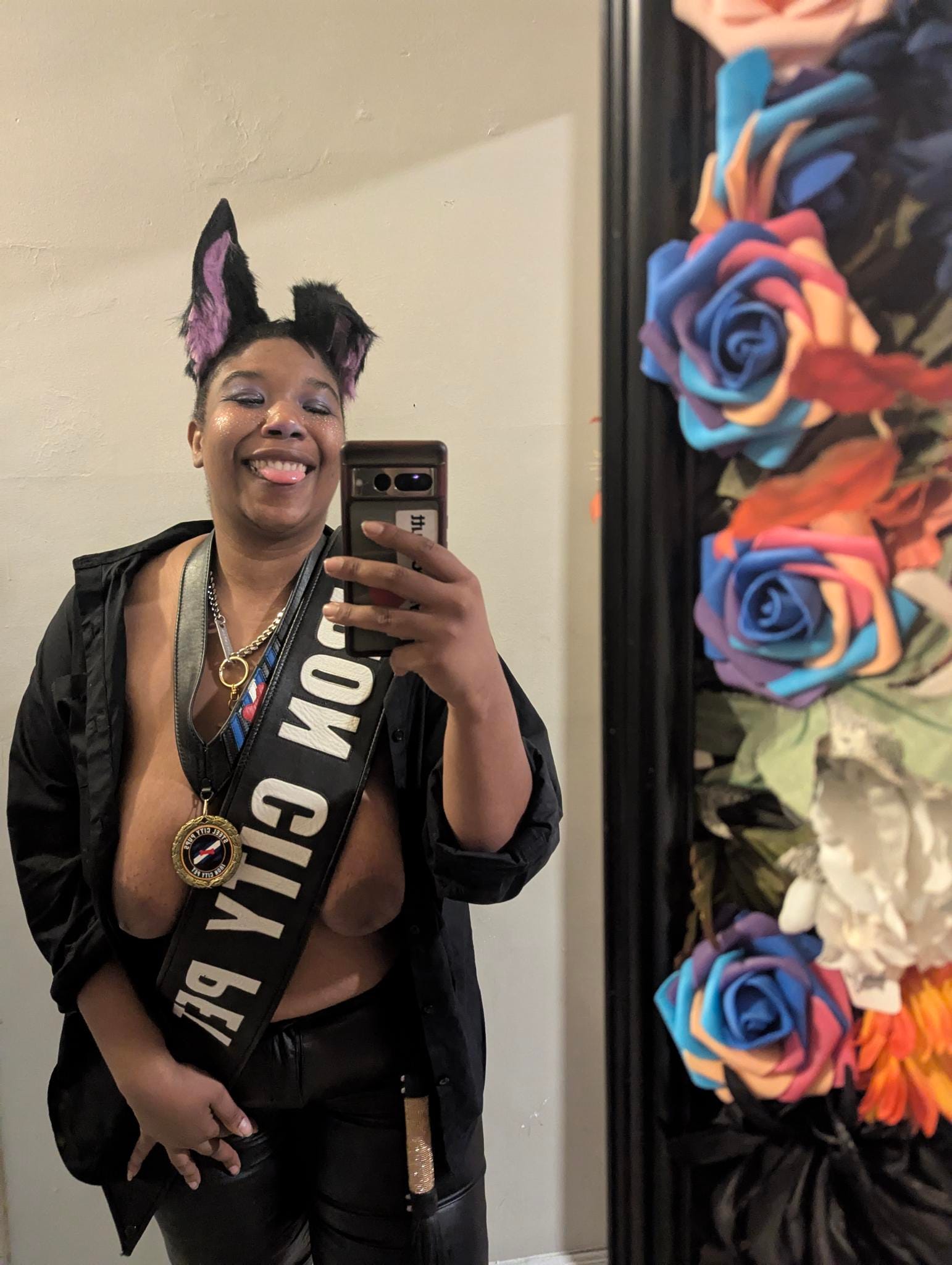 Princess Lavender, a Black pup, smiles for a mirror selfie. They are wearing their "Iron City Pet" leather sash, the "Iron City Pet" medal around their neck, a chain collar, pointed black and purple ears (one perked up and the other bent in half), an open black button-up, and leather pants. A flogger with a shiny handle hangs from their belt loop. They have purple eye makeup on with white freckles dotted on their cheeks. Next to the mirror are colorful roses.