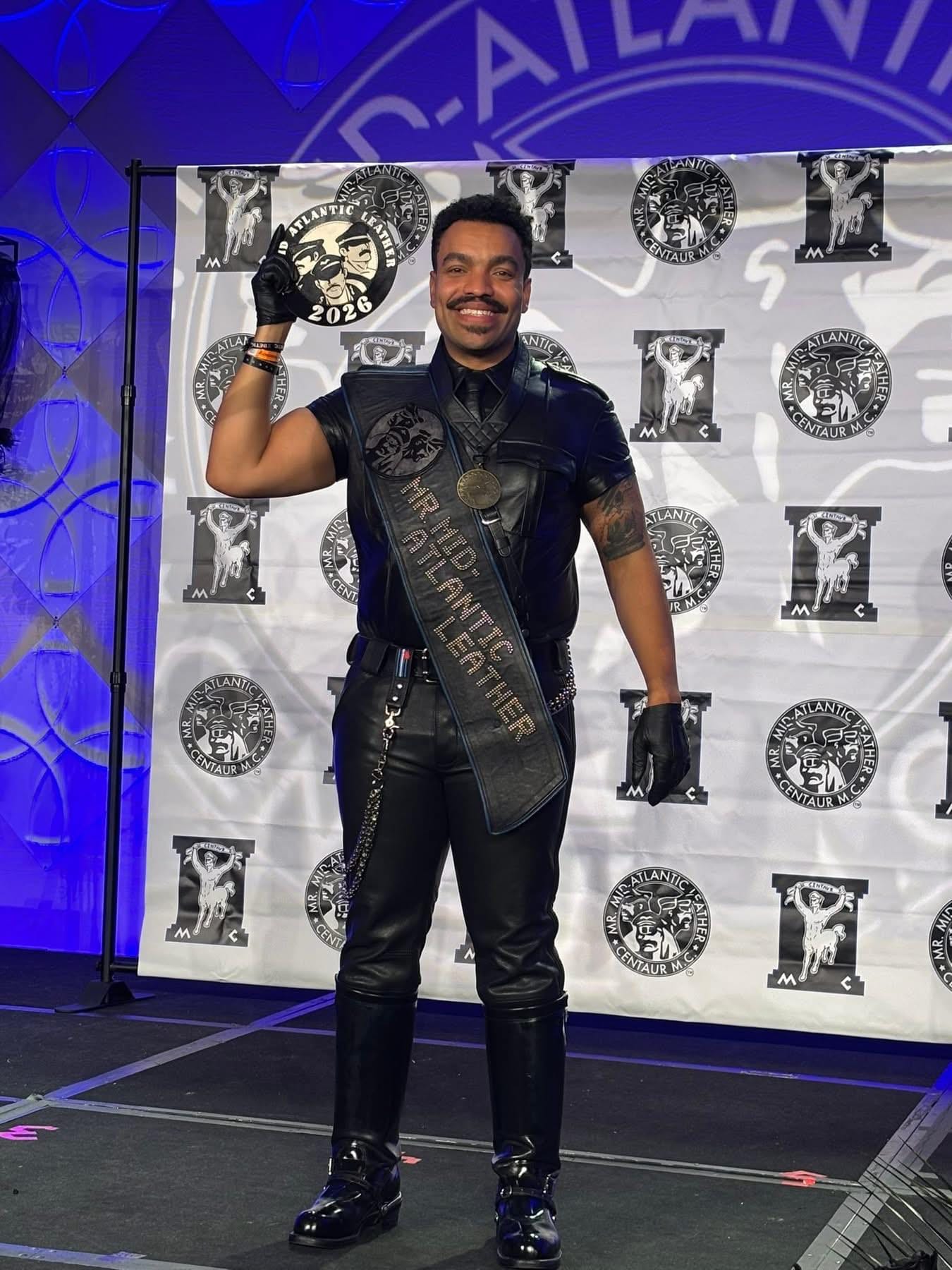 A Black man dressed in full leather, wearing the "Mr. Mid-Atlantic Leather" sash across his chest and holding a circular sign that says "Mr. Mid-Atlantic Leather 2026."
